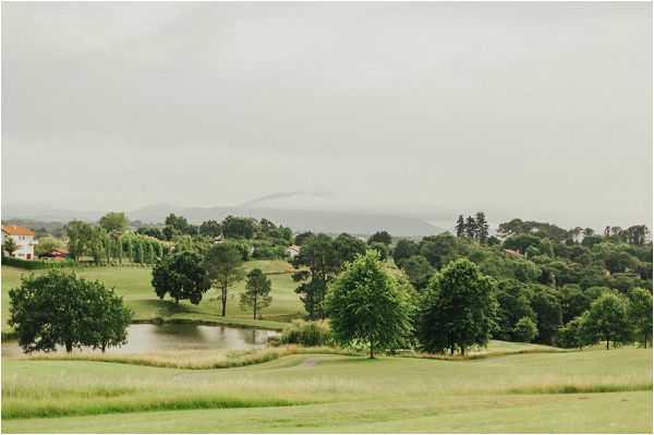 Wide landscape shot of expansive green grounds featuring a small pond, scattered mature trees, and rolling lawns stretching toward a distant treeline and mountain silhouette under an overcast sky. Several white and terracotta-roofed buildings are visible on the left edge of the frame. The image contains no people, wedding party, or decor elements visible. The shot appears to document the venue grounds or surrounding landscape. Potential venue feature image.