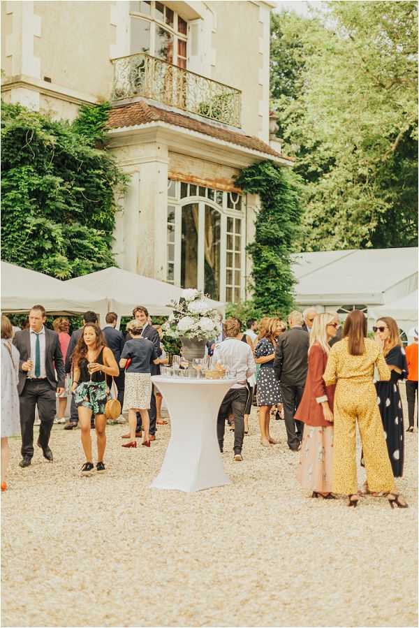 Outdoor cocktail hour taking place on a gravel courtyard in front of a French chateau with ivy-covered stone facade, wrought-iron balcony, and tall arched windows. Approximately 20–30 guests mingle in small groups, dressed in smart-casual summer attire including a yellow printed jumpsuit, floral dress, and polka-dot dress. A white linen cocktail table in the foreground holds glassware and a centerpiece of white blooms, likely hydrangeas or peonies, in a silver vessel. White canopy tents are visible on both sides of the frame, suggesting a larger reception setup. The shot is a medium wide-angle taken at ground level, capturing both the crowd activity and the chateau architecture in the background. Potential venue feature image.