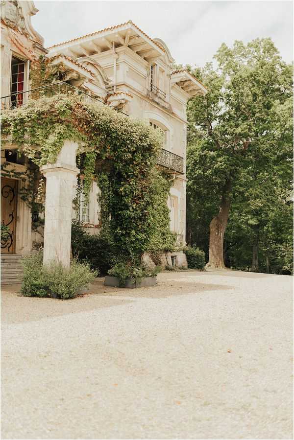 Exterior wide shot of a French château or manor house featuring pale stone architecture with ornate columns, wrought iron balconies, and arched windows heavily draped in climbing ivy. The forecourt is finished with a wide gravel driveway, and low ornamental planters with greenery sit at the base of the columns. No people are visible in the frame. The image has a soft, slightly overexposed tone with a muted color palette. Potential venue feature image.