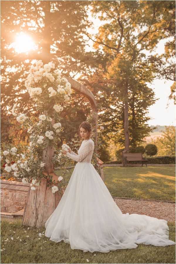A bridal portrait taken outdoors in a garden setting at golden hour, with warm sunlight filtering through the trees behind the subject. The bride stands beside a rustic wooden arch heavily adorned with white climbing roses and green foliage, gently touching the blooms. She wears an ivory ball gown with a long-sleeved, dotted or embellished bodice and a full, flowing tulle skirt with a cathedral-length train spread across the lawn. The styling is romantic and garden-inspired, with a classic-meets-boho aesthetic. The shot is a full-length portrait with a slightly backlit, sun-flare composition.