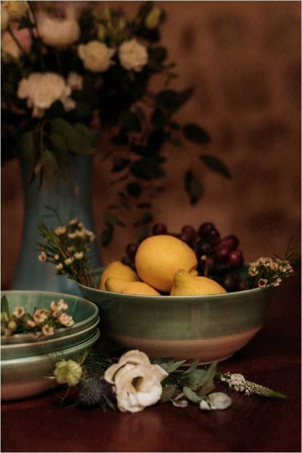 A close-up detail shot of a styled tablescape with a rustic, moody aesthetic. A matte green ceramic bowl holds yellow lemons and dark red grapes, placed on a deep burgundy-toned wooden surface alongside stacked sage-green ceramic plates and a pale blue ceramic vase holding cream roses, eucalyptus, and dark foliage in soft focus in the background. Scattered at the base of the arrangement are loose cream lisianthus blooms, thistle, wax flower sprigs, and eucalyptus leaves, contributing to an earthy, autumn-toned decor palette. The warm, dim lighting and dark background give the composition a painterly, moody quality reminiscent of Dutch still-life styling.