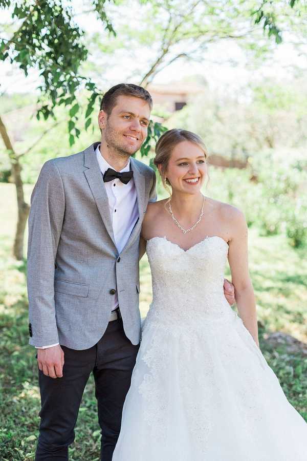 A couples portrait taken outdoors, showing the bride and groom posing together in a lightly wooded garden setting. The groom wears a light grey blazer, dark trousers, a white dress shirt, and a black bow tie, while the bride wears a strapless ivory ballgown with a sweetheart neckline and lace bodice detailing, accessorized with a delicate crystal necklace. The bride's hair is styled in a soft updo and both are smiling directly at the camera. The shot is a mid-length portrait with a shallow depth of field, softly blurring the green foliage and a building structure in the background.