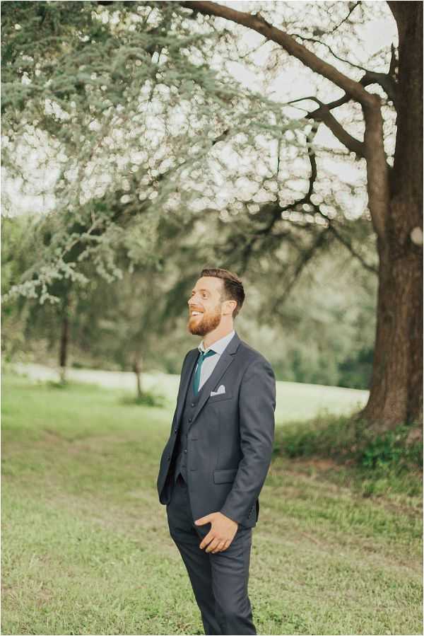 A groom stands outdoors in a garden or parkland setting, smiling and looking upward with one hand in his pocket. He is wearing a charcoal grey three-piece suit with a teal tie and a white pocket square. The portrait is a medium full-length shot with soft, diffused natural light and a blurred green background.