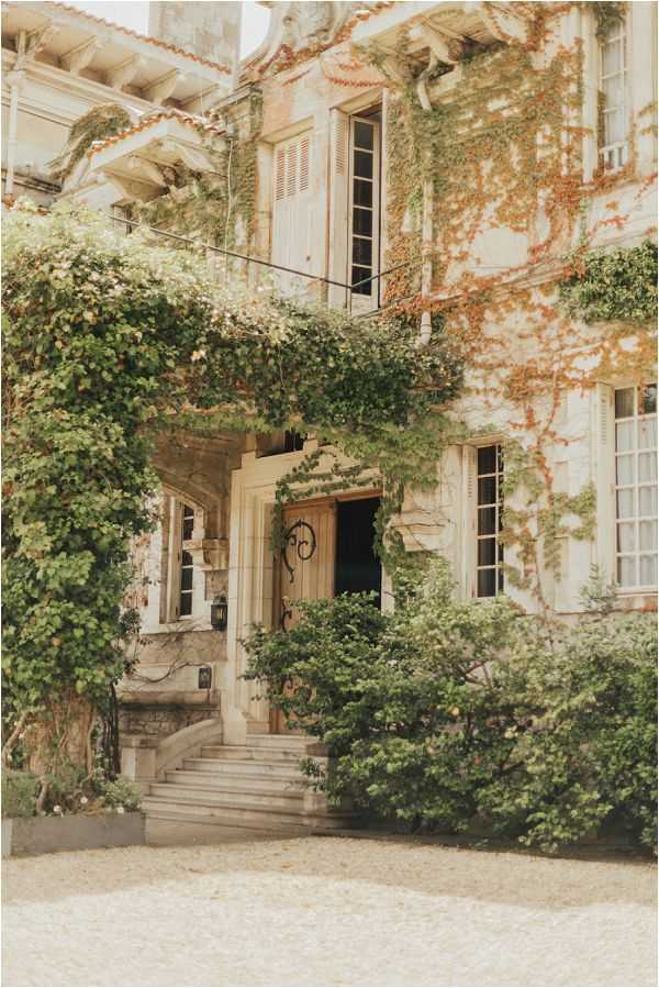 A wide exterior shot of a French manor or chateau facade, showing a multi-story stone building with a warm, aged patina. The entrance features a wooden double door with ornate wrought-iron decorative details, accessed by a set of stone steps flanked by low stone balustrades. The building facade is heavily covered in climbing vines with green and rust-orange foliage, and dense shrubbery lines the base of the structure. A gravel forecourt is visible in the foreground. No people are present in the frame. Potential venue feature image.