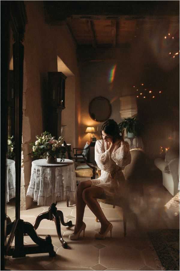 A bride is seated alone in a warmly lit interior room, captured in a contemplative pose with her hands near her face. She is wearing a short white lace bridal robe and nude heels, suggesting this is a getting-ready moment. The room has a classic French country interior with terracotta tile floors, exposed wooden ceiling beams, antique furniture including a wingback chair, and a round side table covered with a white lace cloth holding a floral arrangement of soft pink and white blooms with greenery. Warm fairy lights are visible in the background along with a table lamp, creating a golden ambient glow, and a prismatic light flare crosses the upper portion of the frame as an intentional stylistic element. The shot is a medium full-body portrait taken through or beside a large ornate dark-framed mirror, adding depth to the composition.