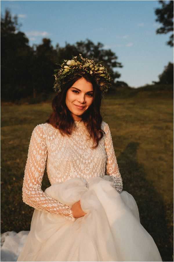 A bridal portrait taken outdoors in a open grassy field during golden hour, with warm late-afternoon light casting a soft glow. The bride wears a white gown with a high-neck, long-sleeved lace bodice featuring a floral crochet pattern, paired with a full tulle skirt that she gathers in her hands. She wears a full flower crown made of greenery, cream buds, and small mixed botanicals in a boho style. The composition is a medium portrait shot with a shallow depth of field, keeping the bride in sharp focus against a softly blurred background of trees and lawn.