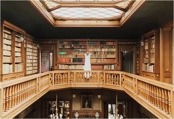 A short white wedding dress is hung from the upper balcony railing of a grand two-story library, centered in the frame against a backdrop of floor-to-ceiling wooden bookshelves filled with leather-bound volumes. The room features warm honey-toned wood paneling, turned wooden balustrade railings on the upper gallery level, and a large skylight overhead providing natural diffused light. Below on the ground floor, a framed portrait painting, decorative vases, and candelabras are visible. The composition is a wide symmetrical shot capturing the full architectural interior of the space. Potential venue feature image.
