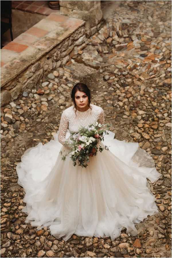 A bridal portrait shot from above, with the bride standing alone outdoors on a cobblestone and pebble ground near rustic stone steps. She is wearing a full-skirted ivory tulle ball gown with a long-sleeved, high-neck lace and dotted illusion bodice, with the skirt spread wide around her. She holds a loose, organic bouquet featuring white blooms, dusty pink flowers, terracotta tones, and trailing greenery and eucalyptus. Her dark hair is pulled back in a low updo. The setting has a rustic, bohemian aesthetic, and the overhead angle emphasizes the dramatic volume of the skirt against the textured ground.