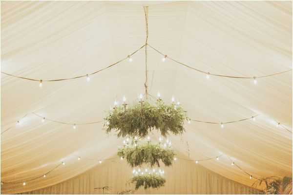 Detail shot of a marquee ceiling decorated with two tiered chandelier-style installations made from lush green foliage, fitted with white candle-style bulbs. Strings of globe fairy lights are draped across the cream-draped tent ceiling, creating a warm, softly lit atmosphere. The overall decor palette is cream and green with a rustic-natural styling theme. No people are visible in the frame.