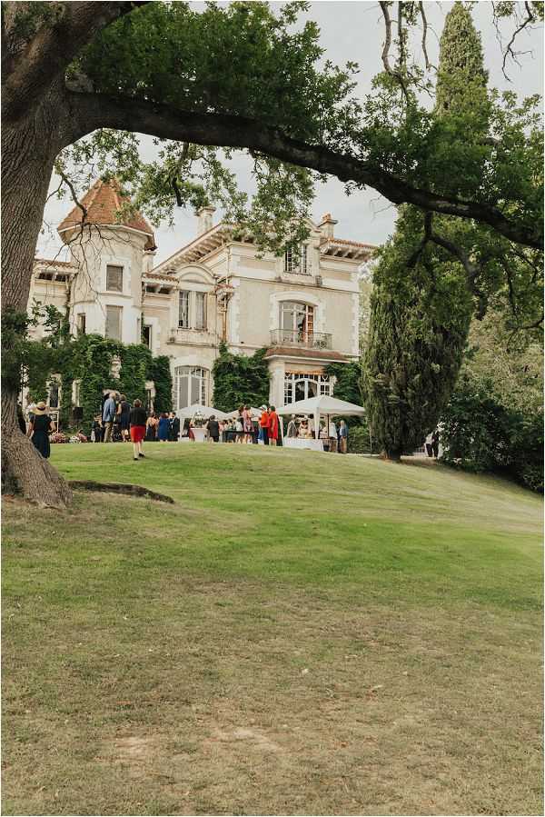 A cocktail hour or outdoor reception gathering is taking place on the grounds of a French chateau, with approximately 20-30 guests mingling in front of the building. The chateau is a large, classic French manor with ivy-covered stone facade, tall arched windows, a balcony, and a distinctive corner turret with an orange-tiled roof. A white umbrella or canopy is set up near the chateau entrance, likely covering a bar or catering station. Guests are dressed in a mix of colorful outfits including a red skirt, blue dress, and various casual-formal attire. The wide shot is framed from a distance through the canopy of a large tree in the foreground, giving a sense of the expansive lawn leading up to the venue. Potential venue feature image.