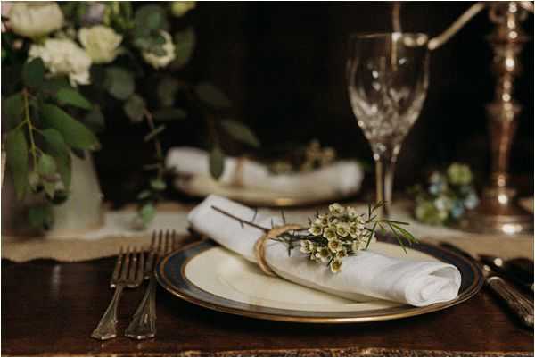 Close-up detail shot of a wedding reception place setting on a dark wood table. The plate stack consists of a cream charger with a dark navy and gold rim, topped with a white linen napkin folded and tied with twine and a small sprig of white waxflower with greenery. Vintage-style silver cutlery is placed on either side, and a cut-crystal wine glass and a silver candlestick are visible in the background. A white ceramic vase with white roses, eucalyptus, and lush greenery is softly out of focus in the background, contributing to a classic, moody tablescape with a dark, warm-toned color palette.