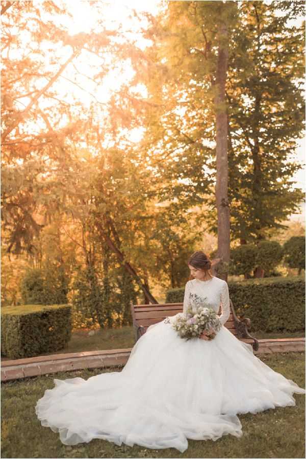 A bridal portrait taken outdoors in a formal garden setting at golden hour, with warm light filtering through the trees. The bride is seated on a dark wood bench, looking downward, wearing a full-ballgown silhouette wedding dress with a long-sleeved lace bodice and a voluminous ivory tulle skirt with an extended train spread across the lawn. She holds a loosely arranged bouquet of soft white, pale lavender, and dusty green blooms, likely including wisteria-style florals and eucalyptus. Her hair is styled in a low updo. The background features neatly trimmed hedgerows and tall trees, and the composition is a medium full-length portrait shot.