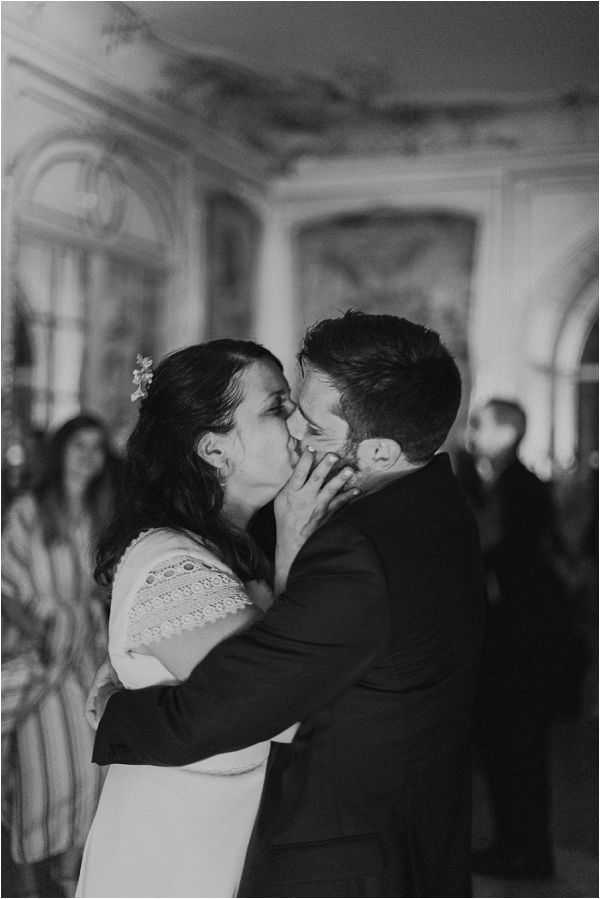A black-and-white close-up portrait of a bride and groom sharing a kiss indoors, likely during a civil ceremony or reception moment. The bride, wearing a white dress with lace detailing on the shoulder and a small floral hair accessory, cups the groom's face with her hand while he wraps his arms around her; the groom is dressed in a dark suit. The background is softly blurred but reveals an ornate interior with decorative molding, painted wall panels, and arched architectural details consistent with a French chateau or hôtel de ville salon, with several guests visible in the background. The image is shot in medium portrait format with shallow depth of field, rendered in high-contrast black and white with deep dark tones in the foreground figures.