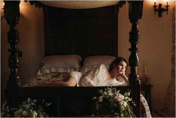 A bride poses in a getting-ready or boudoir-style shot, lying across a large dark wood four-poster bed in a dimly lit interior room. She is wearing a sheer white lace robe and has her hair partially pinned up with loose curls falling around her face. The room has warm amber lighting from wall-mounted candle sconces and a small taper candle on the bedside table, creating a moody, low-light atmosphere. In the foreground, a bridal bouquet featuring soft pink, ivory, and white blooms with greenery is partially visible. The composition is a wide portrait shot taken from a low angle, emphasizing the ornate carved bed frame and the rich, dark interior setting.