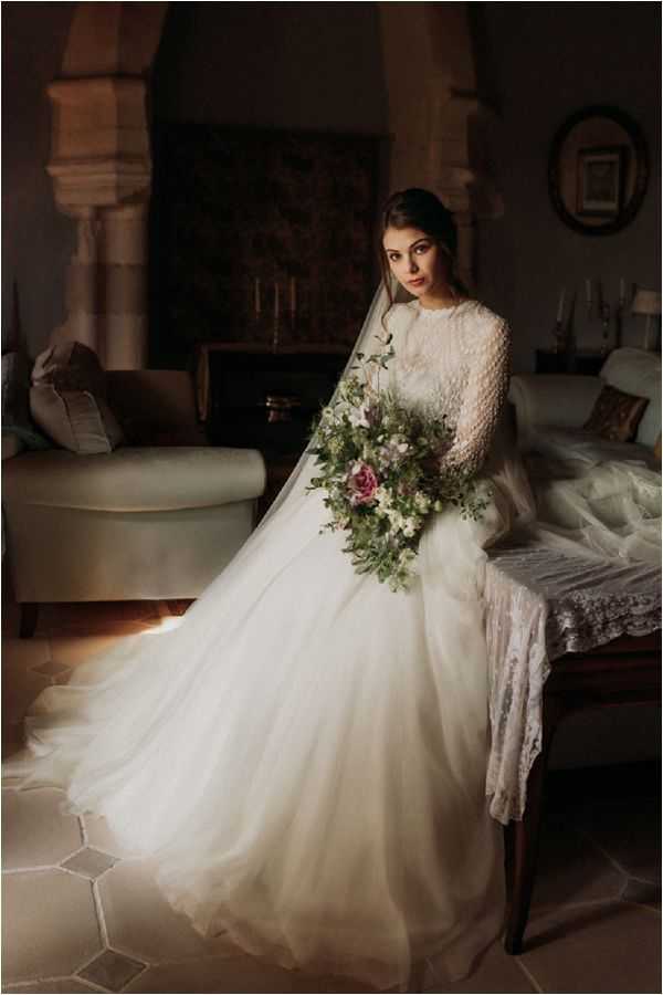 A bridal portrait taken indoors in what appears to be a historic chateau or manor house interior, featuring stone archways, dark wood furniture, and a fireplace with candelabras visible in the background. The bride is seated on a bench at the foot of a bed, wearing a full-skirted ivory ball gown with a long-sleeved lace and pearl-embellished bodice, paired with a long cathedral-length veil draped across her skirt. She holds a loosely arranged cascading bouquet featuring deep pink roses, white blooms, purple wildflowers, and abundant trailing greenery and foliage. The image has a moody, low-key lighting style with warm ambient light illuminating the bride against a dark background, shot as a three-quarter-length portrait with a classic, romantic aesthetic.
