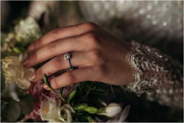 A close-up detail shot of a bride's hand resting on her bridal bouquet, with a diamond halo engagement ring prominently displayed on her ring finger. The bride is wearing a long-sleeved dress with heavily beaded and embroidered lace cuffs in ivory or silver tones. The bouquet beneath her hand includes cream and blush roses, white tulips or freesias, and green foliage, creating a soft, muted floral palette. The image is shot in low, moody lighting with a shallow depth of field that keeps the ring and hand in sharp focus while the background falls into soft bokeh.