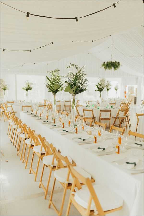 A wide shot of a wedding reception setup inside a white draped marquee tent with no guests present. The space features long banquet tables covered in white linens, set with white plates, white napkins folded with green accents, glassware, and small amber-colored bottles as place settings. Tall glass vase centerpieces hold tropical green palm fronds and large leaves, creating a fresh, botanical aesthetic. Round tables are also visible in the background with similar arrangements. Natural wood folding chairs with white cushioned seats line the tables throughout. Edison bulb string lights hang in loose strands across the white draped ceiling, and suspended greenery wreaths add additional botanical detail overhead. The overall decor palette is white and green with natural wood tones, giving the space a clean, tropical-inspired style.