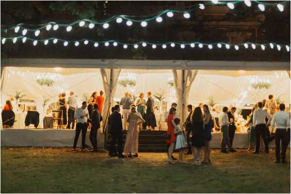 An outdoor evening wedding reception scene showing approximately 30–40 guests gathered around and inside a white open-sided marquee tent. The tent is warmly lit from within, creating a glowing effect against the dark night sky, and strands of globe string lights are strung overhead outside the tent. Guests are dressed in cocktail attire including dark suits, a red dress, a blue dress, and a gold/champagne jumpsuit. The setting appears to be a garden or lawn area. The shot is a wide environmental image taken from a slight distance, capturing both the exterior crowd mingling on the grass and the silhouettes of guests visible through the tent walls.