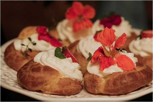 Close-up detail shot of wedding desserts displayed on a white decorative plate. The pastries are cream puffs or choux buns filled with swirled whipped cream and garnished with bright red-orange edible flowers, sliced strawberries, and small fresh herb leaves. The warm, dark background gives the image a moody, candlelit reception atmosphere. The styling suggests a romantic, garden-inspired dessert presentation with a rich color palette of red, cream, and golden brown.
