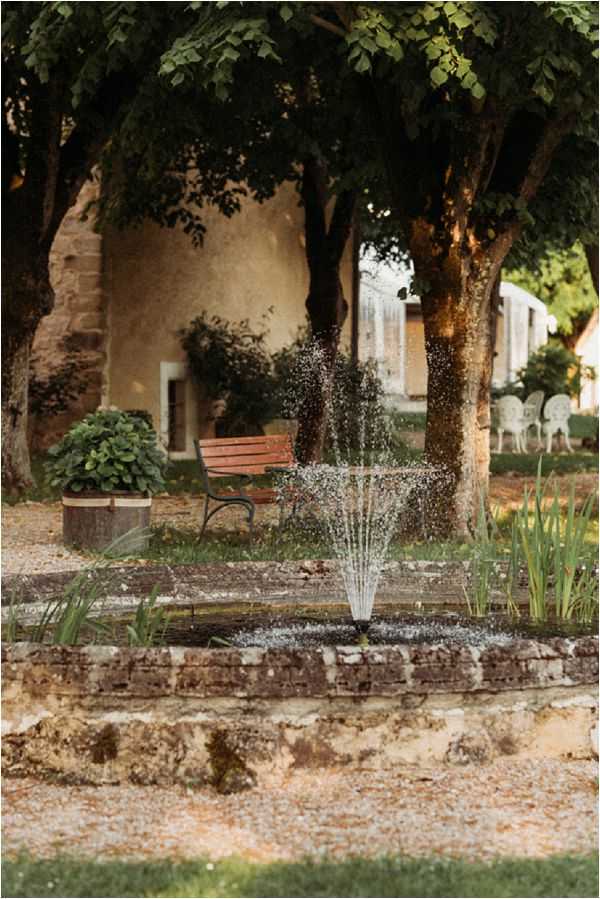 An outdoor garden area at what appears to be a French country estate or chateau, featuring a small stone-edged fountain as the central subject. The grounds include a wooden slatted bench, a large potted plant, and white garden chairs visible in the background to the right. The venue building shows aged ochre-toned rendered walls with a rounded architectural feature. No people are present in this image. The shot is a medium portrait-orientation composition with the fountain in sharp focus and the background softly blurred. Potential venue feature image.