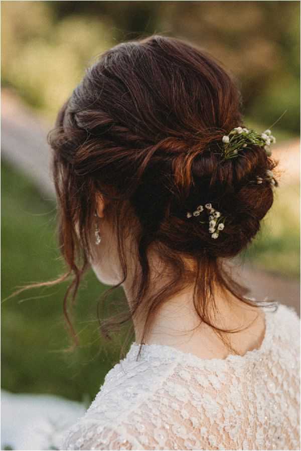 Close-up portrait shot of a bride photographed from behind, showcasing her bridal hair styling. Her dark auburn hair is arranged in a loosely twisted updo with soft, face-framing tendrils left loose around the neck. Small sprigs of white baby's breath are tucked into the bun as the sole hair accessory, alongside what appears to be a small crystal or pearl drop earring visible on her left ear. She is wearing a long-sleeved ivory lace dress with textured floral lace detailing visible at the neckline and sleeves. The background is an outdoor setting with soft, blurred greenery consistent with a garden or natural landscape. The overall styling aesthetic is romantic boho with natural, organic hair accessory choices.