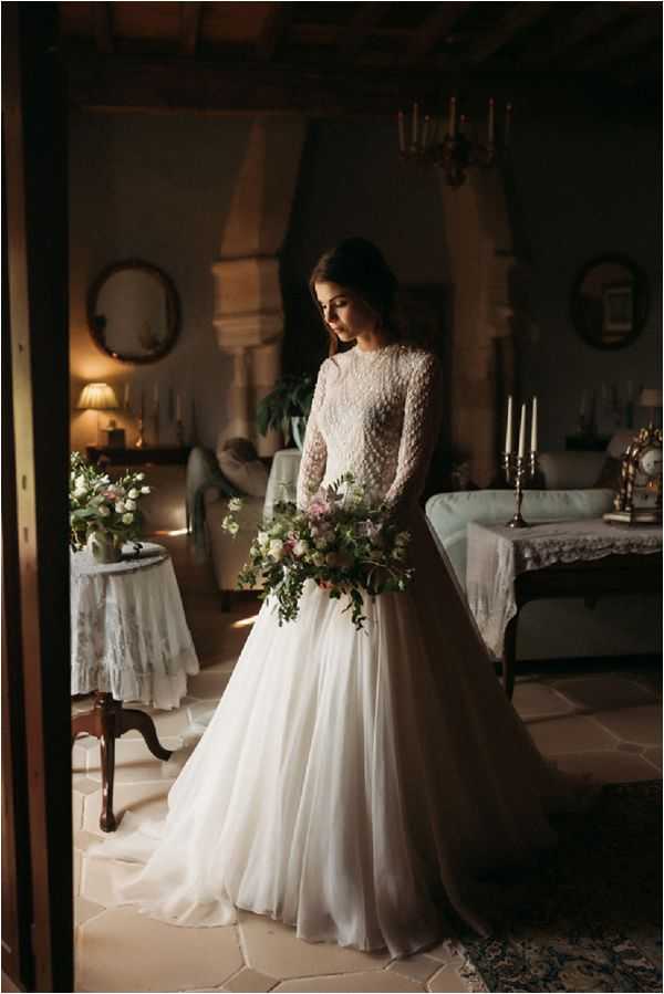 A bridal portrait taken indoors in what appears to be a chateau or manor house interior, with the bride standing in a doorway framed by the dark wooden door surround. The bride wears a long-sleeve white gown with a heavily beaded or pearl-embellished bodice and a flowing chiffon skirt with a train, her dark hair pinned up. She holds a large, loosely arranged bouquet featuring blush and dusty pink roses, white blooms, and abundant trailing greenery. The room behind her features vintage furnishings including a tufted sofa, lace-covered side table with a small floral arrangement, round mirror, candlesticks on a brass candelabra, and warm lamplight creating a moody, low-lit atmosphere consistent with a classic or vintage styling theme. Full-length portrait composition shot from a slight distance, with warm amber tones in the background contrasting against the white of the gown.