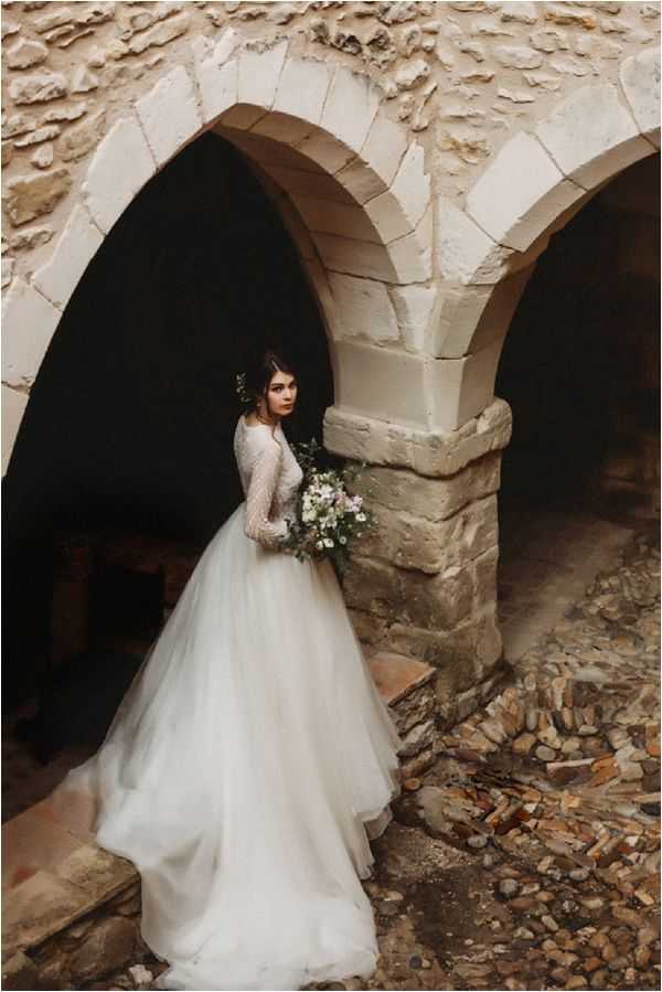 A bridal portrait taken outdoors at what appears to be a historic stone structure with arched doorways, likely a ruined or partially restored chateau or chapel. The bride stands between two stone arches, wearing a full-length white ballgown with long sheer embellished sleeves and a voluminous tulle skirt. She holds a loose, organic bouquet featuring white blooms, small purple flowers, and trailing greenery, and wears a small greenery hair accessory. The composition is a full-length portrait shot with dramatic dark negative space within the arches contrasting against the pale stone architecture and white gown, giving the image a moody, editorial feel with a rustic, romantic styling direction.