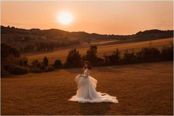 A bridal portrait taken outdoors during golden hour, with the bride standing alone in an open field against a warm orange sunset. The bride wears a white flowing gown with a full tulle skirt that appears to be in motion, creating a sweeping effect across the ground, paired with a long-sleeved lace bodice. She is turned slightly away from the camera, looking back over her shoulder toward the horizon. The wide shot captures a rolling countryside landscape with hills in the background, bathed in deep amber and orange sunset light. The overall styling is romantic and airy, consistent with a boho or soft classic aesthetic.