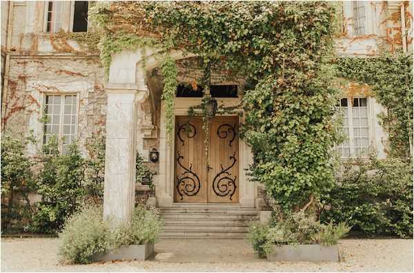 Wide shot of the exterior entrance of a French chateau or manor house, showing no people. The focal point is a set of large double wooden doors with ornate black wrought-iron scrollwork detailing, approached by a short flight of stone steps. The stone facade is heavily covered in climbing green vines and foliage, and large rectangular planters flank the entrance steps. The overall palette is warm amber, green, and aged stone. No wedding party or decor elements are visible beyond the architectural setting. Potential venue feature image.
