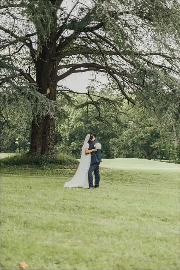 A couple portrait taken outdoors on the grounds of what appears to be a château or golf estate, with the bride and groom embracing beneath a large mature cedar tree. The bride wears a white gown with short sleeves and a long flowing veil, holding a round white bouquet, while the groom is dressed in a navy suit. The shot is a wide full-length portrait, positioning the couple small within the frame to emphasize the expansive landscaped grounds and the dramatic spreading branches of the tree above them. The overall color palette is soft and muted with warm natural light filtering through the foliage.