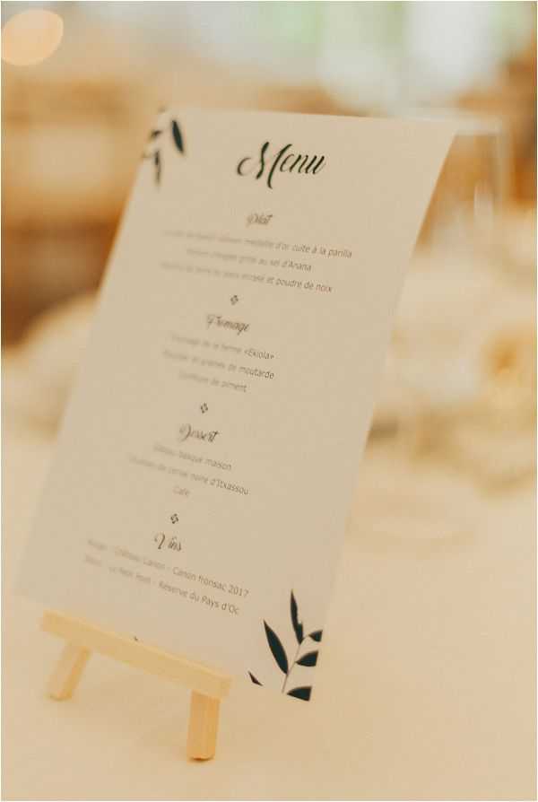 Close-up detail shot of a French wedding reception menu card displayed on a small wooden easel on a white linen-covered table. The menu card is white with dark navy or forest green botanical leaf illustrations in the corners and features calligraphy-style script headings in French, listing courses including Plat, Fromage, Dessert, and Vins, with wine selections including a Canon Fronsac 2017 and Réserve du Pays d'Oc. The background is softly blurred, showing glassware and warm ambient lighting consistent with an indoor reception setting. The overall stationery style is minimal and modern with a botanical accent theme.