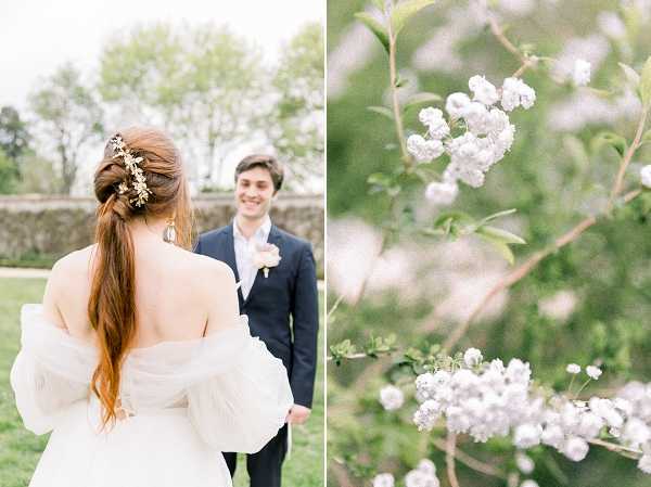 A two-panel image featuring an outdoor portrait and a botanical detail shot. In the left panel, the bride and groom face each other in a garden setting; the bride is seen from behind wearing an off-the-shoulder white gown with voluminous sheer puff sleeves and a low open back, her auburn hair styled in a low ponytail adorned with a gold floral hair piece, while the groom faces the camera smiling, dressed in a navy suit with a white boutonniere. The right panel is a close-up, softly focused detail shot of small white clustered blossoms on a branch with green leaves, shot with shallow depth of field. The overall aesthetic is light, airy, and romantic, consistent with a classic French garden wedding style.