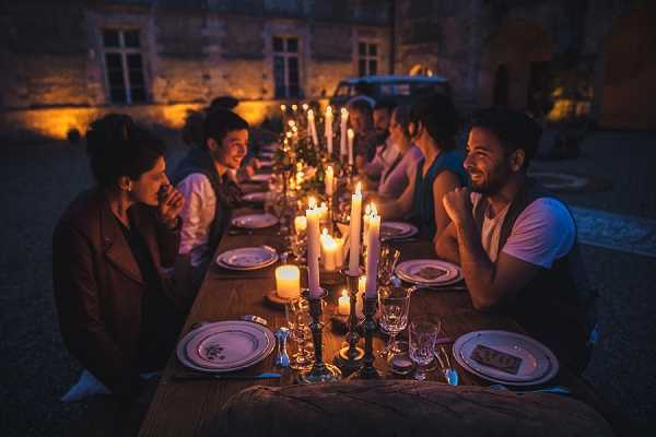 An outdoor evening reception dinner set in a château courtyard, with approximately eight to ten guests seated along a long rustic wooden table lit entirely by candlelight from multiple taper candles in dark metal candlestick holders. The table is set with white china plates, crystal glassware, and scattered greenery as a centerpiece, creating a warm amber glow against the dark night. Guests are dressed in smart-casual attire including waistcoats and blazers, and are engaged in lively conversation; a vintage car and the warmly lit château facade are visible in the background. The composition is a wide-angle shot taken from one end of the table, emphasizing the length of the table and the candlelit atmosphere.