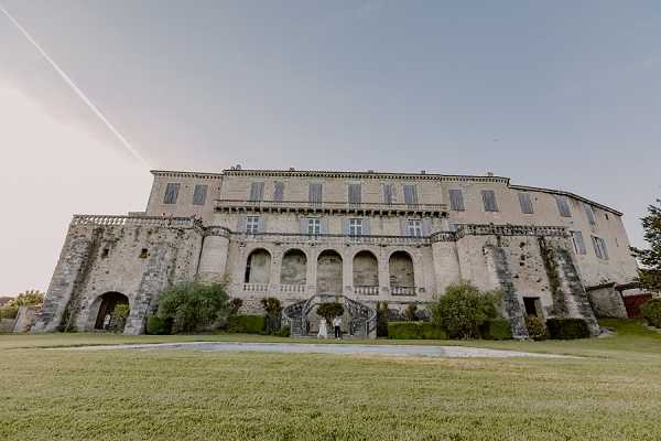 A wide-angle exterior shot of a large French château featuring stone masonry, arched ground-floor loggias, a grand double staircase, and upper-floor shuttered windows with balustrade detailing. A couple in wedding attire — the bride in a white gown and the groom in a dark suit — stand at the base of the central staircase, appearing very small against the scale of the building. The grounds in the foreground are manicured lawn with trimmed hedges flanking the façade. The composition is a low-angle wide shot that emphasizes the architecture over the subjects. Potential venue feature image.