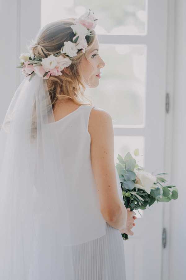 A bridal portrait shot from behind and to the side, taken indoors near a bright white-framed window. The bride wears a sleeveless, minimalist white gown with a pleated skirt and a long tulle veil, topped with a flower crown featuring blush pink and white lisianthus blooms with greenery. She holds a small bouquet of white lisianthus and eucalyptus at her back, gazing toward the window in profile. The overall styling is soft and romantic-boho with a light, airy palette of white, blush, and pale green.