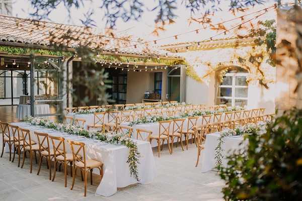 An outdoor wedding reception setup in a stone courtyard of what appears to be a French mas or bastide property. The tables are arranged in long banquet-style rows covered with white linen tablecloths, lined with natural wood cross-back chairs. The table runners feature lush greenery garlands with small white florals, likely eucalyptus and white blooms, cascading down the center. Warm Edison bulb string lights are strung overhead in a grid pattern across the courtyard, adding ambient lighting. The wide-angle shot captures the full table layout from a slightly elevated perspective, framed by out-of-focus foliage in the foreground. The overall decor palette is white and green with natural wood tones, consistent with a classic rustic-chic styling. Potential venue feature image.