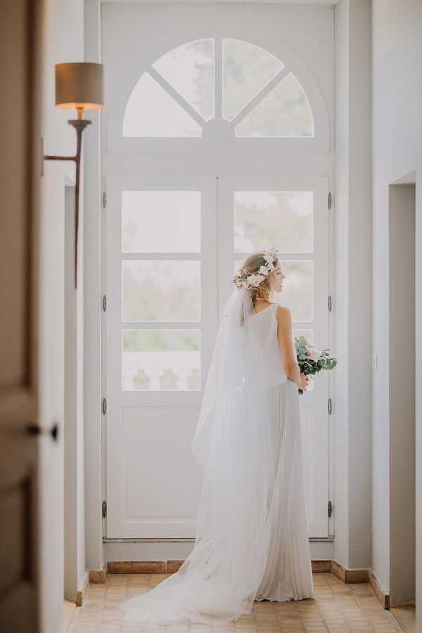 A bridal portrait taken indoors, showing the bride standing with her back three-quarters to the camera in front of a tall white arched double door with fanlight window. She is wearing a sleeveless ivory pleated gown with a long cathedral-length veil and a floral hair crown featuring soft white and blush blooms. She holds a small bouquet with white flowers and greenery. The interior space has white walls, terracotta tile flooring, and a wall-mounted lamp with a gold shade visible to the left. The overall styling is romantic and boho-leaning, with natural light flooding through the door. Full-length portrait composition.