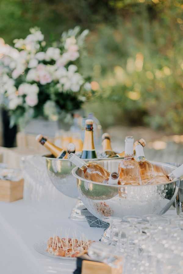 An outdoor cocktail hour beverage station featuring two large polished silver ice buckets filled with chilled rosé and champagne bottles. The table is covered with a white linen and set with crystal glassware and small appetizers on toothpicks arranged on a plate in the foreground. A floral arrangement of blush pink and white blooms is visible in soft focus in the background, alongside additional bottles on wooden crates. The overall decor palette is white, silver, and blush, suggesting a classic, light-toned styling. Close-up detail shot with shallow depth of field.