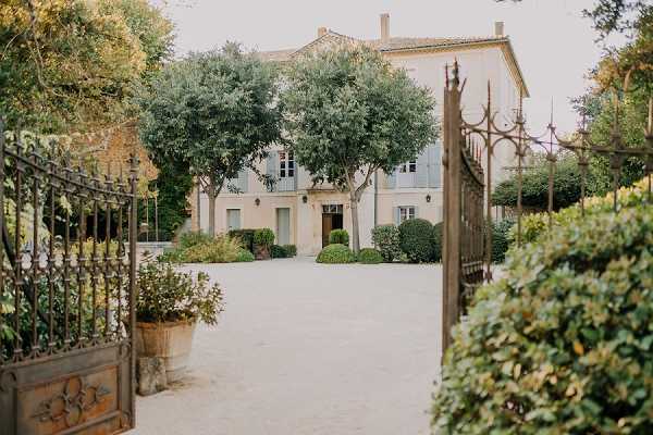 Wide shot of a classic French manor house or bastide photographed through open ornate wrought-iron gates, with a gravel courtyard leading to the main entrance. The two-story stone building features pale ochre-toned render, blue-grey shuttered windows, and a central wooden door flanked by mature olive or plane trees and clipped boxwood shrubs. Large terracotta pots with greenery frame the gate entrance in the foreground. No people are visible; this is a venue exterior shot taken in natural daylight with a soft, muted palette. Potential venue feature image.