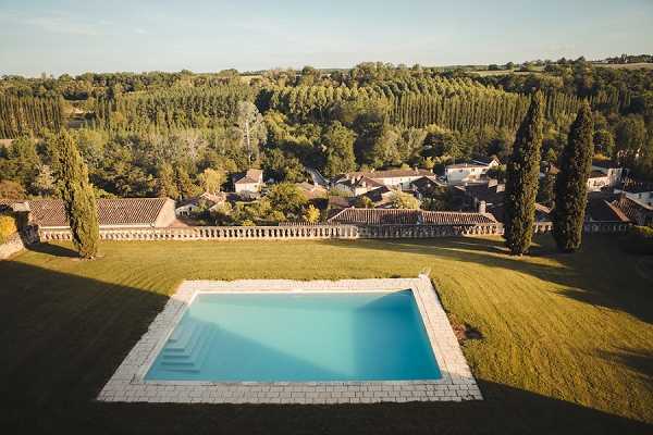 Aerial wide shot of a French country estate's outdoor grounds, featuring a rectangular stone-edged swimming pool with blue water on a large lawn. The property includes a balustraded stone terrace wall, terracotta-roofed outbuildings, and tall cypress trees flanking the grounds. The surrounding landscape shows dense woodland and what appears to be a small village in the distance. No people are visible in this image. Potential venue feature image.