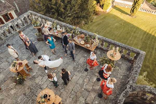 An aerial wide shot of a cocktail hour taking place on a stone terrace of what appears to be a French chateau or manor house, with a well-manicured lawn visible beyond a balustrade in the background. Approximately 15–20 guests are mingling across the terrace, with the bride visible in a white dress and veil near the center of the frame and other guests dressed in a mix of casual and semi-formal attire including a teal dress and an orange skirt. Small round wooden bistro tables are scattered across the terrace, and a long wooden food or drinks table is set up along one side, styled with rustic wooden elements, bowls, and natural decor. The overall styling is relaxed and rustic, with warm late-afternoon sunlight casting long shadows across the cobblestone terrace. Potential venue feature image.