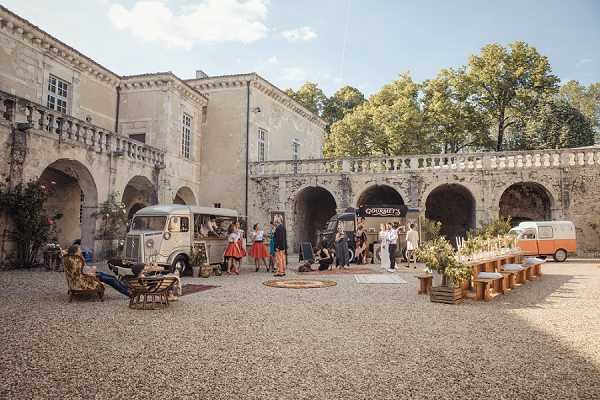 A cocktail hour or outdoor reception setup in the graveled courtyard of a historic French chateau with stone arched colonnades and a balustrade. Two vintage vehicles serve as food and drink stations — a grey Citroën HY van and an orange VW camper van — with a sign reading 'Gourmet's' visible on one of the arches. Approximately 15–20 guests mingle in the courtyard, with some seated in rattan chairs and others standing in small groups. A long wooden banquet table lined with wooden benches runs along the right side, decorated with tall greenery and floral arrangements in warm tones, with wooden crate accents. The styling has a rustic, bohemian aesthetic with warm earth tones. Wide establishing shot capturing the full courtyard scene. Potential venue feature image.