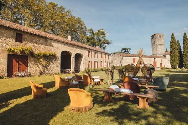 Outdoor cocktail hour or lounge area set up on a lawn in front of a stone farmhouse or domaine with arched openings and terracotta roof tiles, with a stone tower visible in the background. The seating arrangement features mustard yellow velvet armchairs with fringe detailing, rust-orange and teal upholstered chairs, and low wooden rustic bench tables with white bowl-shaped decorative pieces. A bohemian-style teepee or rattan hanging chair structure is visible mid-ground, along with wicker accent chairs and small cocktail tables scattered across the lawn. The styling palette leans heavily boho-rustic with warm earth tones — ochre, terracotta, and teal — and two white bicycles are parked near the arched facade on the left. Wide establishing shot capturing both the decor setup and the full venue exterior. Potential venue feature image.