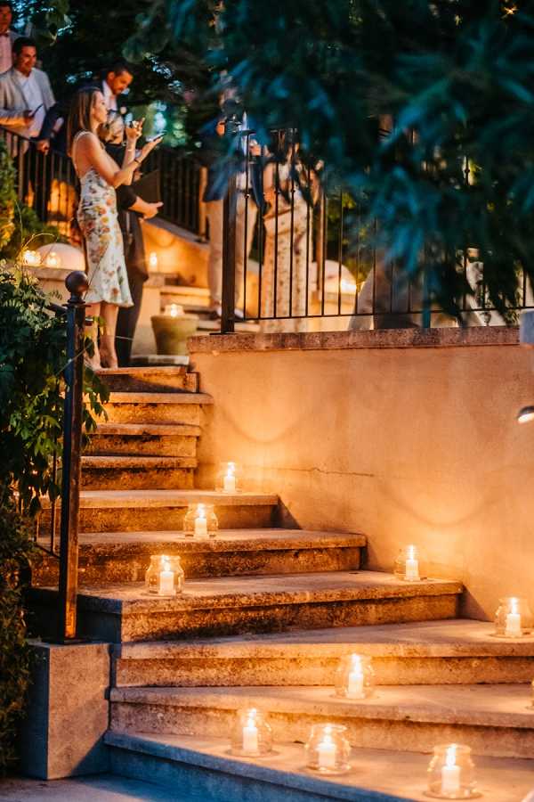 An evening outdoor reception scene at what appears to be a chateau or villa, with wide stone steps lined with small glass votive candles casting a warm amber glow across each step. Several guests are visible at the top of the staircase, including a woman in a white floral-print midi dress who appears to be photographing with her phone. The warm candlelight against the stone architecture creates a rich contrast with the dark blue evening sky and surrounding greenery. This is a medium wide shot emphasizing the candlelit staircase decor detail with guests mingling in the background. Potential venue feature image.