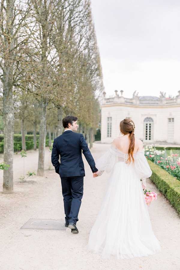 A couple portrait shot from behind as the bride and groom walk hand-in-hand along a gravel path in a formal French garden. The groom wears a navy suit with dark dress shoes and glances back over his shoulder, while the bride wears an off-the-shoulder white tulle gown with voluminous detached sleeves and a low open back, her auburn hair styled in a half-up arrangement with a small gold hair accessory. She carries a bouquet of pink peonies. The background features a classical French pavilion with white stone facade, arched windows, and ornamental statuary along the roofline, flanked by rows of bare-branched trees and manicured hedgerows with pink flowering plants. The composition is a wide portrait shot with a soft, airy light quality. Potential venue feature image.