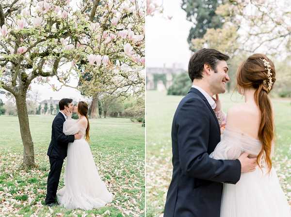 A two-panel portrait of a couple during outdoor wedding portraits on the grounds of what appears to be a French château or manor house. In the left panel, the couple shares a kiss beneath a magnolia tree in full pink bloom, with fallen petals on the grass and the manor building visible in the background. In the right panel, a closer portrait shows the couple facing each other and smiling, with the bride's back to the camera revealing a low-back, off-the-shoulder blush tulle ballgown and an updo decorated with a delicate gold floral hair accessory; the groom wears a dark navy suit. The styling is classic and romantic, with the blush tulle dress complementing the soft pink magnolia blossoms. Both panels are airy, light-filled medium-distance and close portrait shots with a soft, bright exposure.