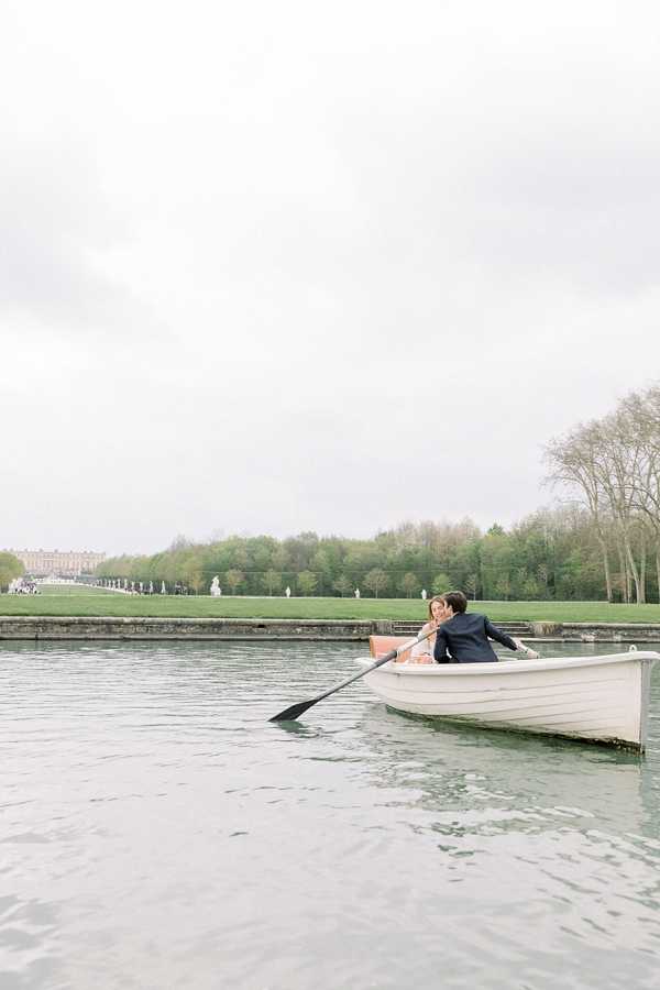 A couple portrait session taking place on the Grand Canal at the Palace of Versailles, with the groom rowing a white wooden rowboat while the bride leans in close to him. The groom is dressed in a navy suit and the bride appears to be wearing a light-colored, likely blush or ivory, wedding dress. The wide shot is taken from water level, capturing the full length of the boat on the canal, with the formal gardens, classical statues, and the Versailles palace building visible in the distant background under an overcast sky. Potential venue feature image.