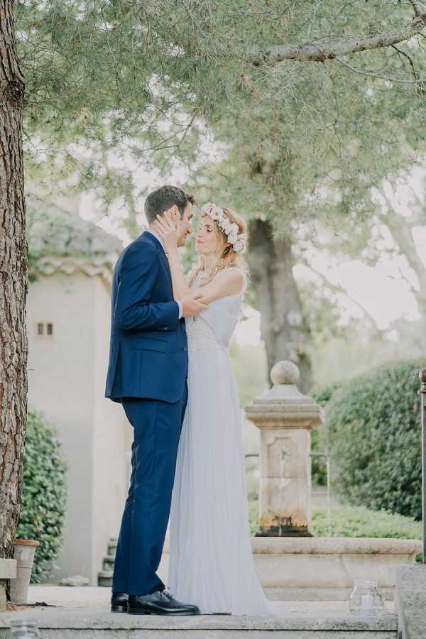 A couple shares an intimate moment outdoors in a Provençal garden setting, their foreheads nearly touching in a quiet embrace. The groom wears a navy blue suit with a white shirt, while the bride wears a flowing white pleated gown with a lace bodice and a floral crown featuring white and blush daisy-like blooms in her loose blonde hair. The styling has a relaxed, boho feel. In the background, a stone fountain or garden ornament is visible alongside trimmed hedges and a white-rendered building with terracotta roof tiles. The shot is a full-length portrait with soft, diffused natural light giving the image a light, airy quality.
