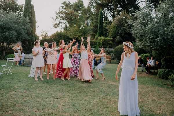 The bouquet toss is taking place outdoors in a garden setting with manicured lawns, cypress trees, and decorative hedging. The bride stands in the foreground on the right, wearing a pale blue pleated two-piece gown and a floral crown with white blooms, her back turned as she has just released the bouquet. A group of approximately twelve female guests reaches and jumps to catch it, dressed in a variety of colorful outfits including a red floral maxi dress, blush pink dress, bright red dress, and other mixed patterns and pastels. Additional seated guests are visible in the background near a wooden bench and white folding chairs. The shot is a wide, candid action frame taken during what appears to be an outdoor cocktail hour or reception in a Provençal-style garden.