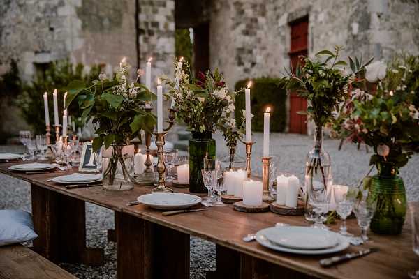 A styled outdoor reception tablescape set on a rustic wooden farm table with bench seating, photographed in a wide-angle detail shot at dusk. The table is decorated with a mix of white taper candles in brass candlesticks, large white pillar candles on wooden slice risers, and floral arrangements in dark green glass bottles and vases featuring lush greenery, deep burgundy blooms, white flowers, and trailing foliage. Place settings include white plates layered on chargers with vintage-style cut crystal glassware. The overall decor palette combines brass, dark green, burgundy, and natural wood tones in a romantic, rustic-organic style. The setting is an outdoor courtyard of a stone chateau or castle, with a red wooden door and weathered stone walls visible in the background. Potential venue feature image.